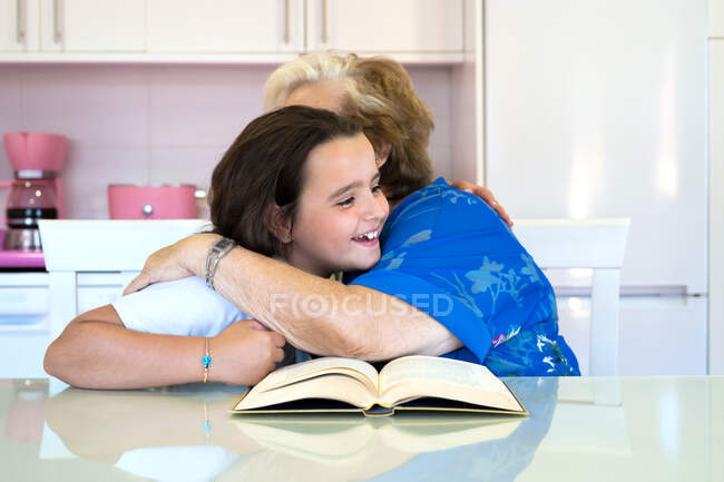 Seitenansicht des fröhlichen Mädchens umarmt bis zur Unkenntlichkeit ältere Großmutter, während sie am Tisch sitzt und tagsüber gemeinsam in der hellen Küche Bücher liest — Stockfoto