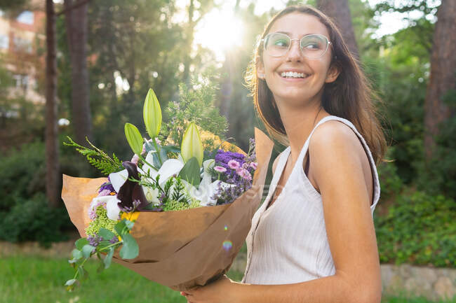 Vue latérale d'une jeune femme sincère dans des lunettes avec un bouquet floral en fleurs regardant loin dans le dos éclairé — Photo de stock