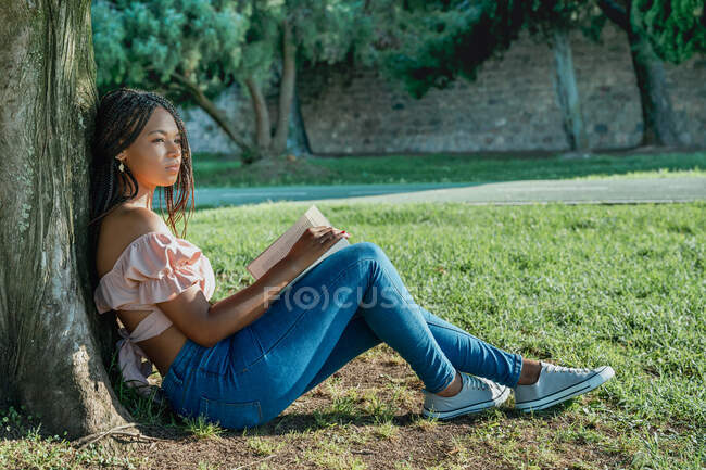 Seitenansicht einer verträumten jungen schwarzen Frau mit Lehrbuch und Afro-Zöpfen, die wegschaut, während sie mit überkreuzten Beinen auf dem Rasen sitzt — Stockfoto