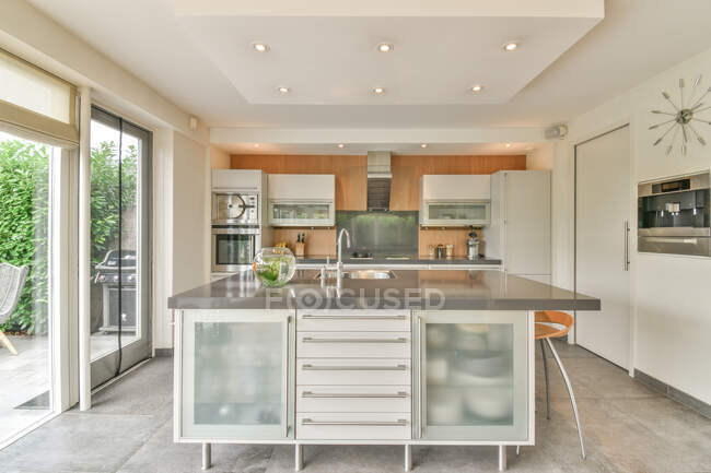 Moderne Kücheneinrichtung mit Tisch und Hockern gegen Kühlschrank und Schränke im Haus mit Glaswand tagsüber — Stockfoto