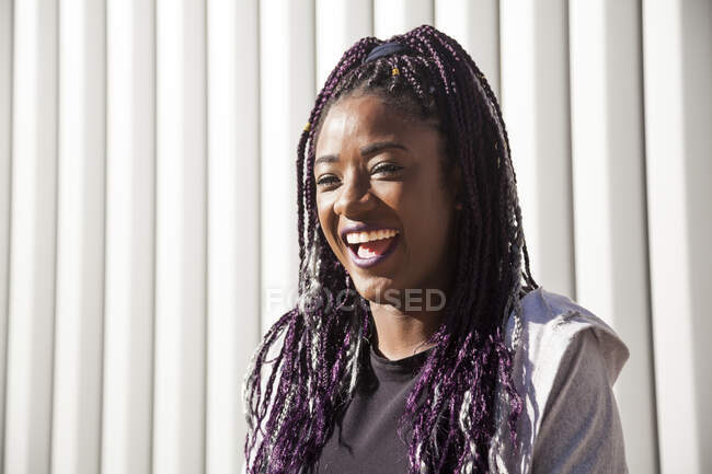 Giovane donna afroamericana felice con lunghe trecce tinte che sorridono brillantemente e distolgono lo sguardo divertendosi contro il muro grigio alla luce del sole — Foto stock