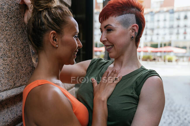 Vista laterale della giovane coppia lesbica allegra alla moda con tatuaggio che si abbraccia guardandosi in un momento di bacio appoggiato a un muro in città — Foto stock