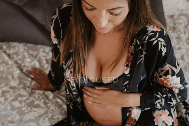 Dall'alto bella donna incinta in biancheria intima seduta guardando giù sulla morbida copertina del letto a casa — Foto stock