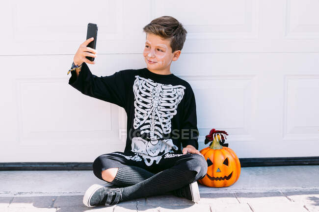 Smiling boy in black skeleton costume with painted face sitting near carved Halloween pumpkin and taking selfie on smartphone near white wall on street — Stock Photo