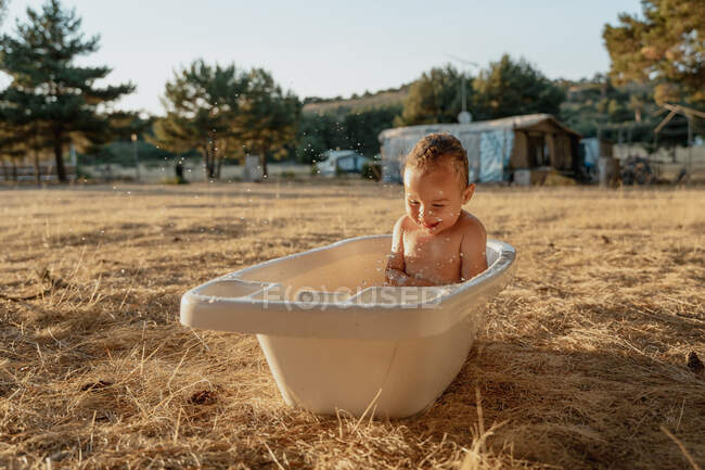 Счастливый ребенок с игрушкой, сидящий в пластиковой ванне, играя с водой в сельской местности — стоковое фото
