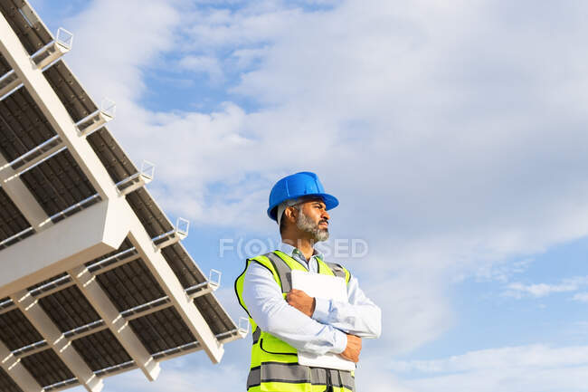 Bajo ángulo de madura étnica inspector masculino en chaleco y hardhat con portapapeles mirando hacia otro lado mientras está de pie cerca de la estación de energía solar - foto de stock