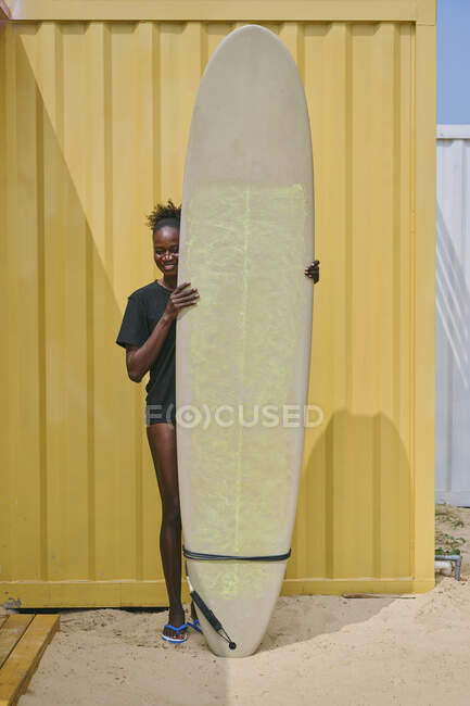 Cheerful young African American sportswoman in bikini and t shirt looking at camera with surfboard in a yellow container on the coast — Stock Photo