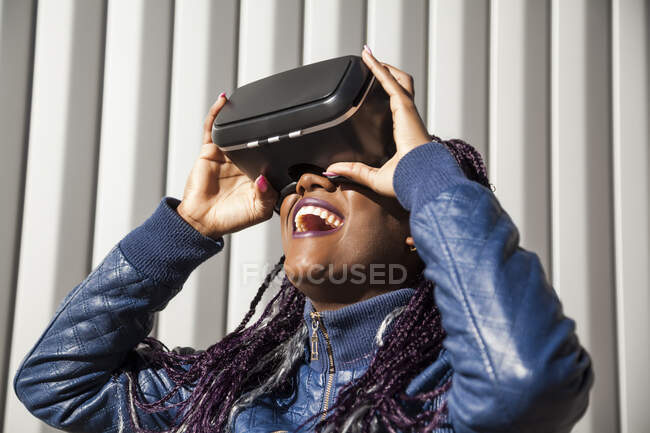 Jovem afro-americana excitada no fone de ouvido VR entretendo e jogando jogo virtual contra a parede listrada cinza — Fotografia de Stock