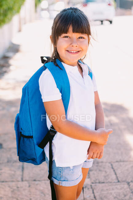 Joyeux écolier aux cheveux bruns en t-shirt blanc et avec sac à dos coloré regardant la caméra sur une passerelle carrelée en ville — Photo de stock