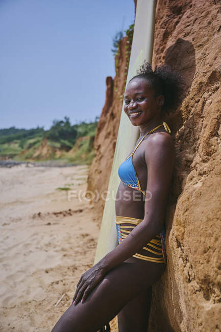 Вид сбоку на афроамериканскую спортсменку, смотрящую на камеру с доской для серфинга со стороны пляжа и перед когтистой скалой — стоковое фото