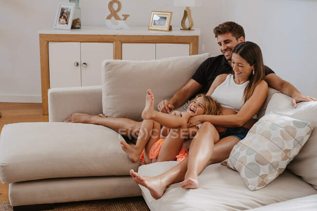 Беззаботный ребенок играет с беременной матерью и улыбается папе во время отдыха на диване в гостиной — стоковое фото