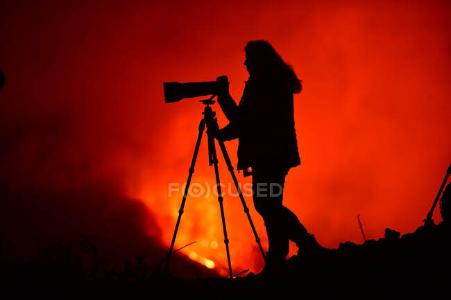 Vista lateral de la silueta de una mujer fotografiando con un teleobjetivo y trípode la explosión de lava en La Palma Islas Canarias 2021 - foto de stock
