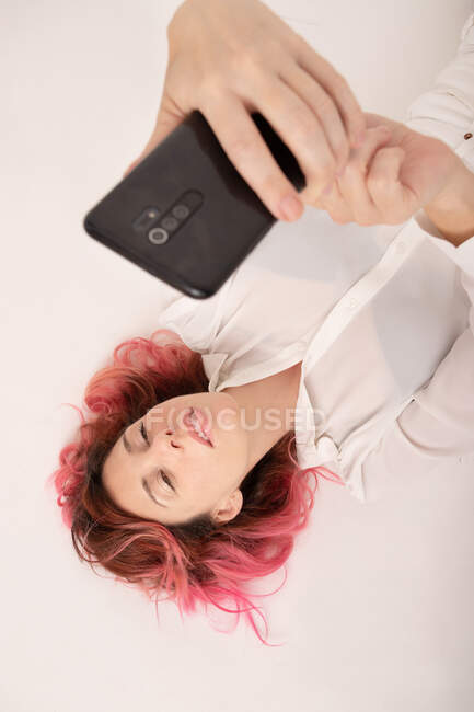 Von oben eine charmante Frau mit pinkfarbenen Haaren in weißer Bluse, die im hellen Raum liegt und ein Selfie mit dem Smartphone macht — Stockfoto