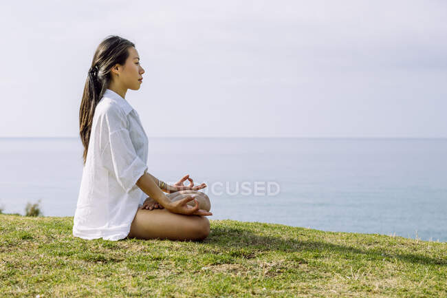 Vue latérale de la jeune femme ethnique rêveuse avec les yeux fermés méditant à Padmasana pose tout en pratiquant le yoga sur la côte de la mer — Photo de stock