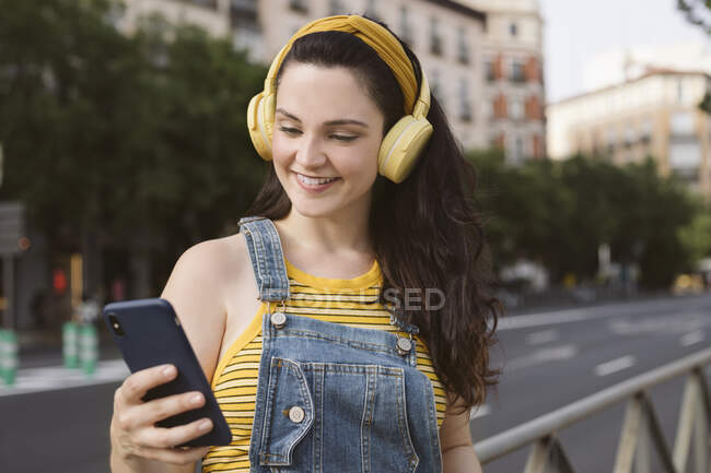 Безтурботна жінка в бездротових навушниках, що переглядають смартфон на вулиці — стокове фото
