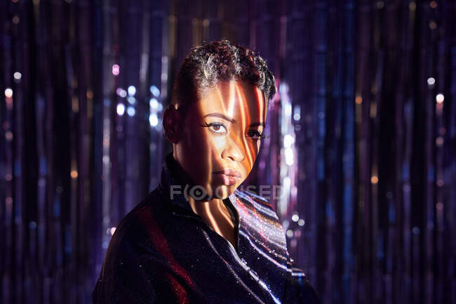 Jeune femme afro-américaine à la mode avec des cheveux courts et des nuances sur le visage en regardant la caméra dans les rayons de lumière — Photo de stock