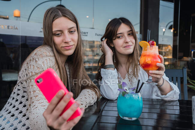Las mejores amigas con vasos de bebidas refrescantes tomando autorretrato en el teléfono celular en la mesa en la cafetería urbana - foto de stock