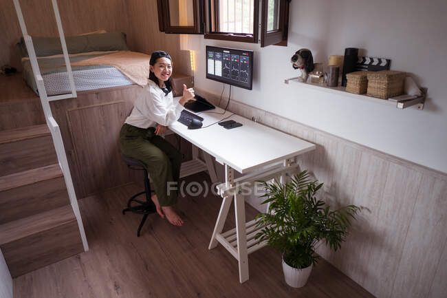 Sonriente corredor étnico femenino escribiendo en el teclado contra el monitor con gráficos y mostrando como gesto mientras mira a la cámara en casa - foto de stock