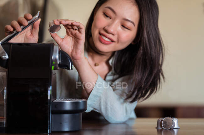 Урожай усміхнений молода етнічна жінка кладе кавовий стручок у виробника на стіл на домашній кухні — стокове фото