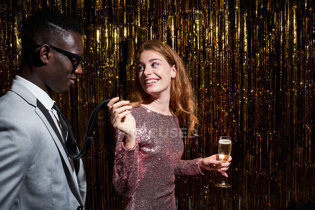 Улыбающаяся женщина с бокалом шампанского держит галстук черного парня, взаимодействуя и глядя друг на друга в канун Нового года — стоковое фото