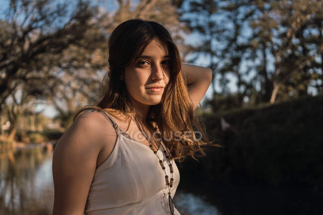 Sanfte Teenagerin in Sundress und Perlen, die Haare berühren, während sie im weichen Sonnenlicht gegen den Fluss in die Kamera blickt — Stockfoto