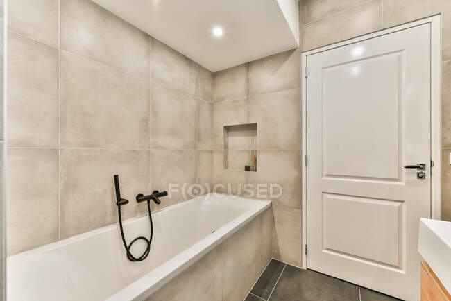 Интерьер стильной ванной комнаты с глянцевой дверью рядом с раковиной и ванной с душем в черепичной стене под огнями в потолке — стоковое фото