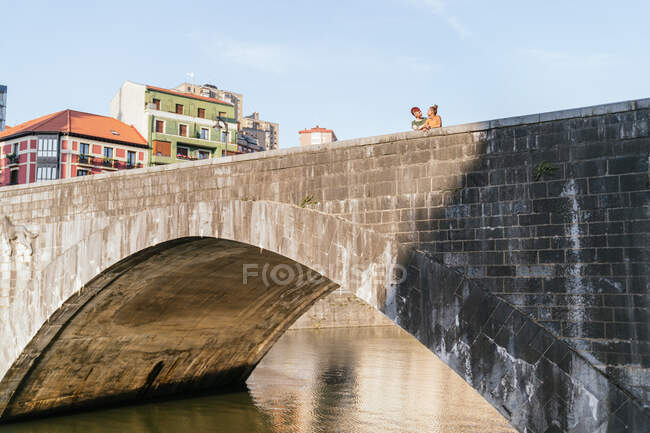 Гомосексуальні жінки говорять і дивляться один на одного на арочному кам'яному пішохідному мосту над водяним каналом під хмарним блакитним небом на сонячному світлі — стокове фото