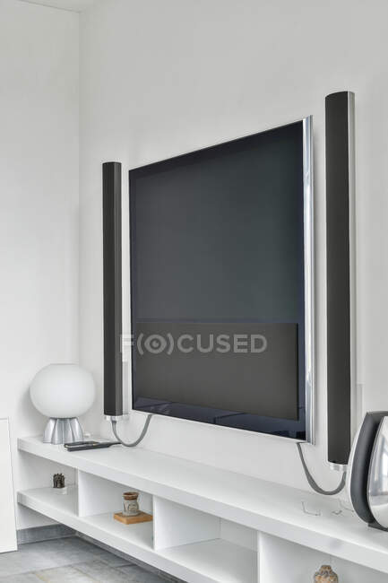 Zeitgenössischer Fernseher mit akustischen Lautsprechern, die in einer minimalistischen Wohnung unter Regalen an der Wand hängen — Stockfoto