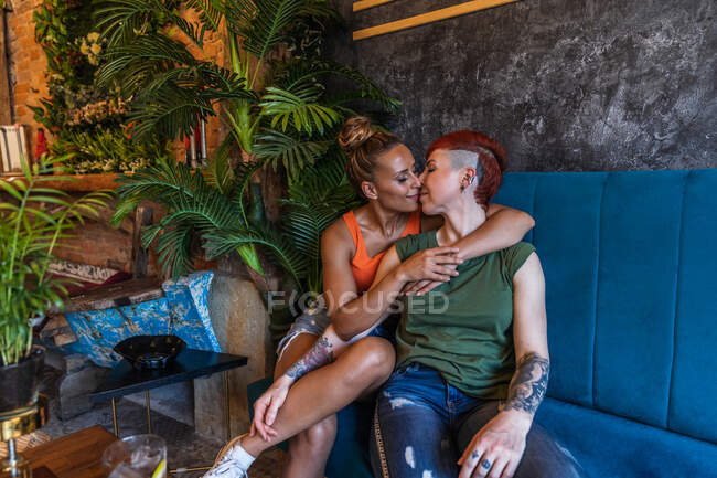 Contenido joven mujer tatuada con mohawk y bebida abrazando y besando a novia lesbiana mientras se miran en el sofá en casa - foto de stock