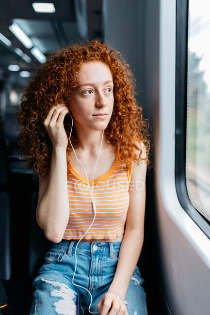 Giovane donna candida con i capelli rossi ricci e il cellulare che ascolta la canzone dagli auricolari mentre guarda lontano in treno — Foto stock