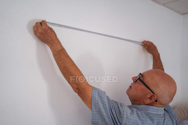 Back view of focused senior male in glasses with raised arms measuring white wall with tape while looking up in house — Stock Photo