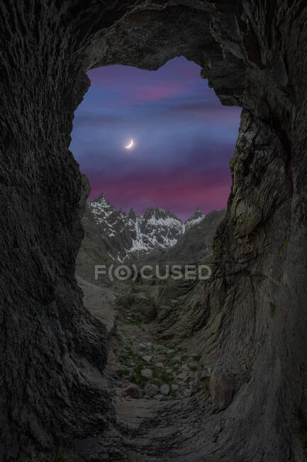 Vista desde la cueva contra formaciones rocosas nevadas bajo el cielo nublado en el crepúsculo con luna creciente - foto de stock