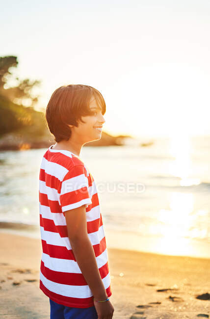 Seitenansicht eines nachdenklichen Jungen, der am nassen Sandstrand steht, der bei Sonnenuntergang vom wogenden blauen Meer gewaschen wird — Stockfoto