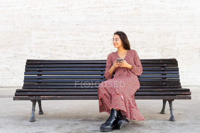 Corpo inteiro de fêmea positiva em roupas elegantes navegando smartphone e sentado em banco de madeira na rua durante o dia — Fotografia de Stock