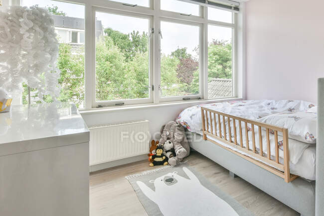 Juguetes colocados en el suelo con alfombra cerca de una cómoda cama en el dormitorio de los niños con gran ventana durante el día - foto de stock