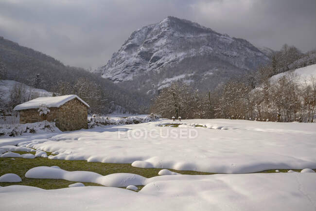 Paesaggio paesaggio di pendii di montagna e valle coperti da neve bianca con piccola casa rurale sotto il cielo nuvoloso blu nel Parco Naturale di Redes situato in Caleao Asturie Spagna — Foto stock