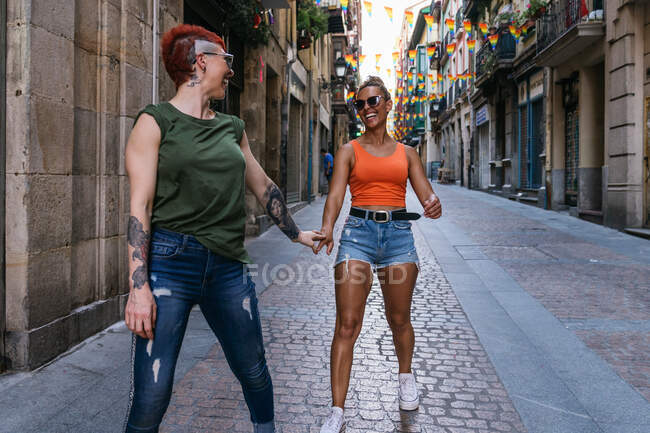Vista lateral de la feliz pareja femenina homosexual joven con tatuajes en ropa de moda mirándose mientras se divierten en la calle - foto de stock