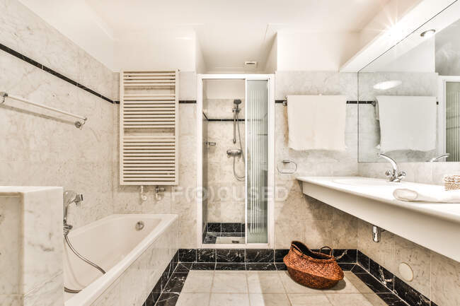 Интерьер современной белой кафельной ванной комнаты с ванной и душевой кабиной возле раковины под зеркалом и полотенцем — стоковое фото