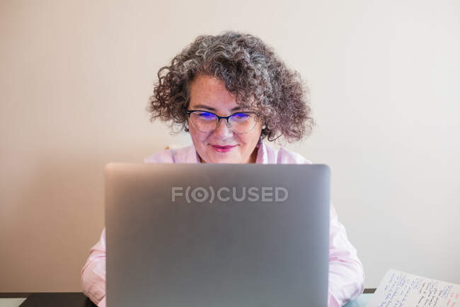 Smiling senior businesswoman in eyewear and earphones sitting at table with netbook — Stock Photo