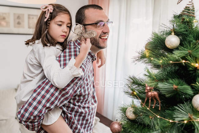 Pai dando porquinho de volta passeio para a filha decorar abeto de Natal com estrela festiva enquanto se prepara para a celebração — Fotografia de Stock