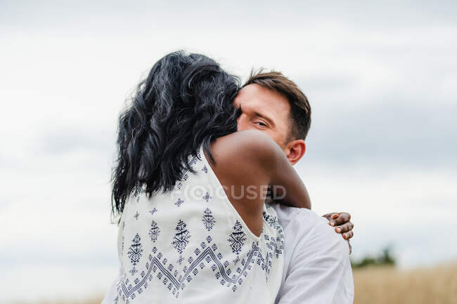 Felice uomo abbracciando fidanzata indiana in piedi in campo sotto cielo nuvoloso — Foto stock