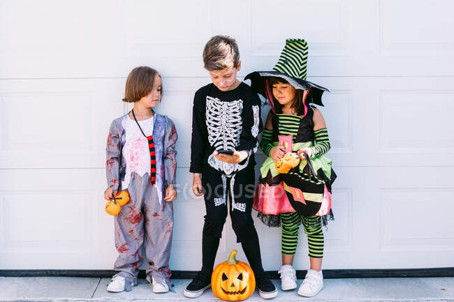 Полное тело группы маленьких детей, одетых в различные костюмы на Хэллоуин с вырезанным Джеком О Фонарем, просматривающих мобильный телефон вместе, стоя рядом с белой стеной на улице — стоковое фото