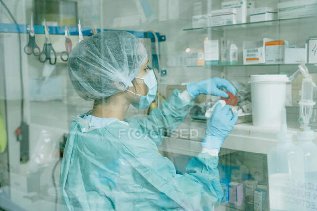 Através da vista lateral da parede de vidro do veterinário fêmea no uniforme médico que toma o rolo adesivo do remendo da prateleira no hospital — Fotografia de Stock