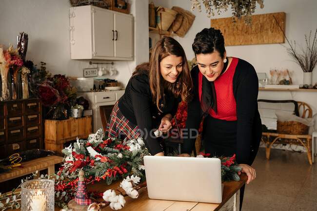 Positive Floristinnen stehen mit Laptop am Tisch und basteln Weihnachtsdekoration im stilvollen Floristikstudio — Stockfoto
