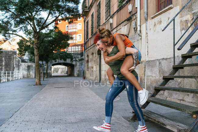 Positive lesbian female giving cheerful beloved piggyback ride while having fun near staircase and urban buildings — Stock Photo