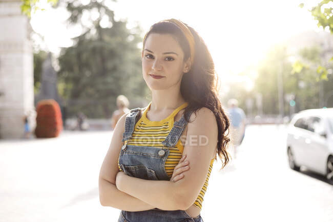 Young female with long brown hair wearing denim overall standing with folded hands and looking at camera with smile — Stock Photo
