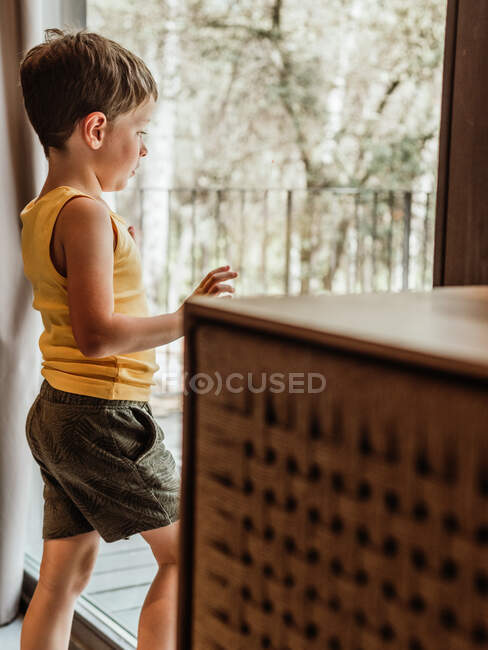 Heiteres kleines Kind steht am Fenster in Hütte und schaut in Gedanken weg — Stockfoto