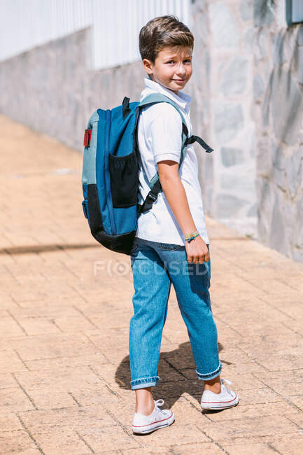 Schulkind in zerrissenen Jeans und Gummischuhen schaut über die Schulter auf gefliestem Bürgersteig in sonniger Stadt — Stockfoto
