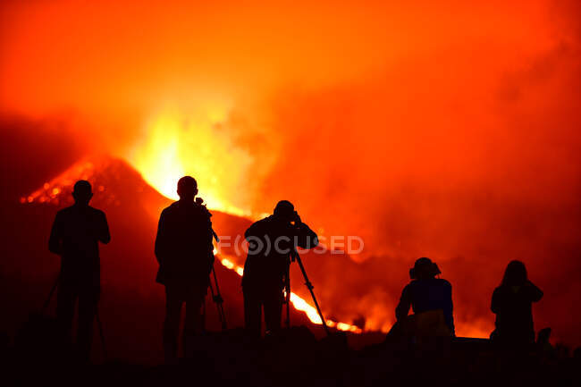 Human silhouettes standing recording and photographing with tripods the exploding lava in La Palma Canary Islands 2021 and two seated silhouettes observing the natural phenomenon. — Stock Photo