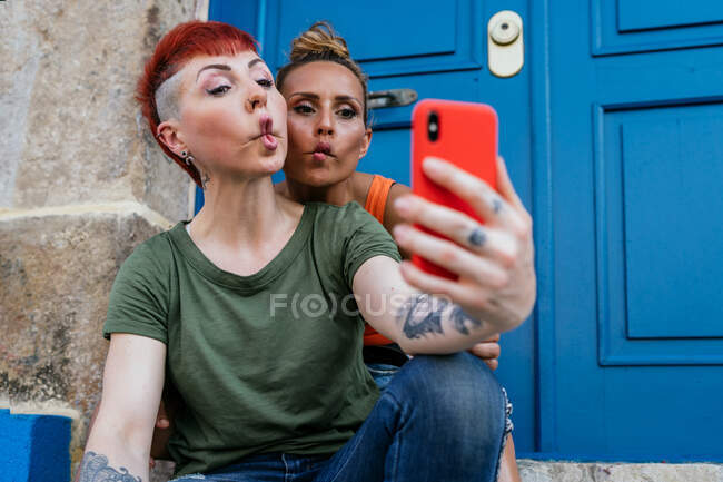 Femmes homosexuelles avec des tatouages grimaçant tout en prenant autoportrait sur téléphone portable contre la porte d'entrée en ville — Photo de stock